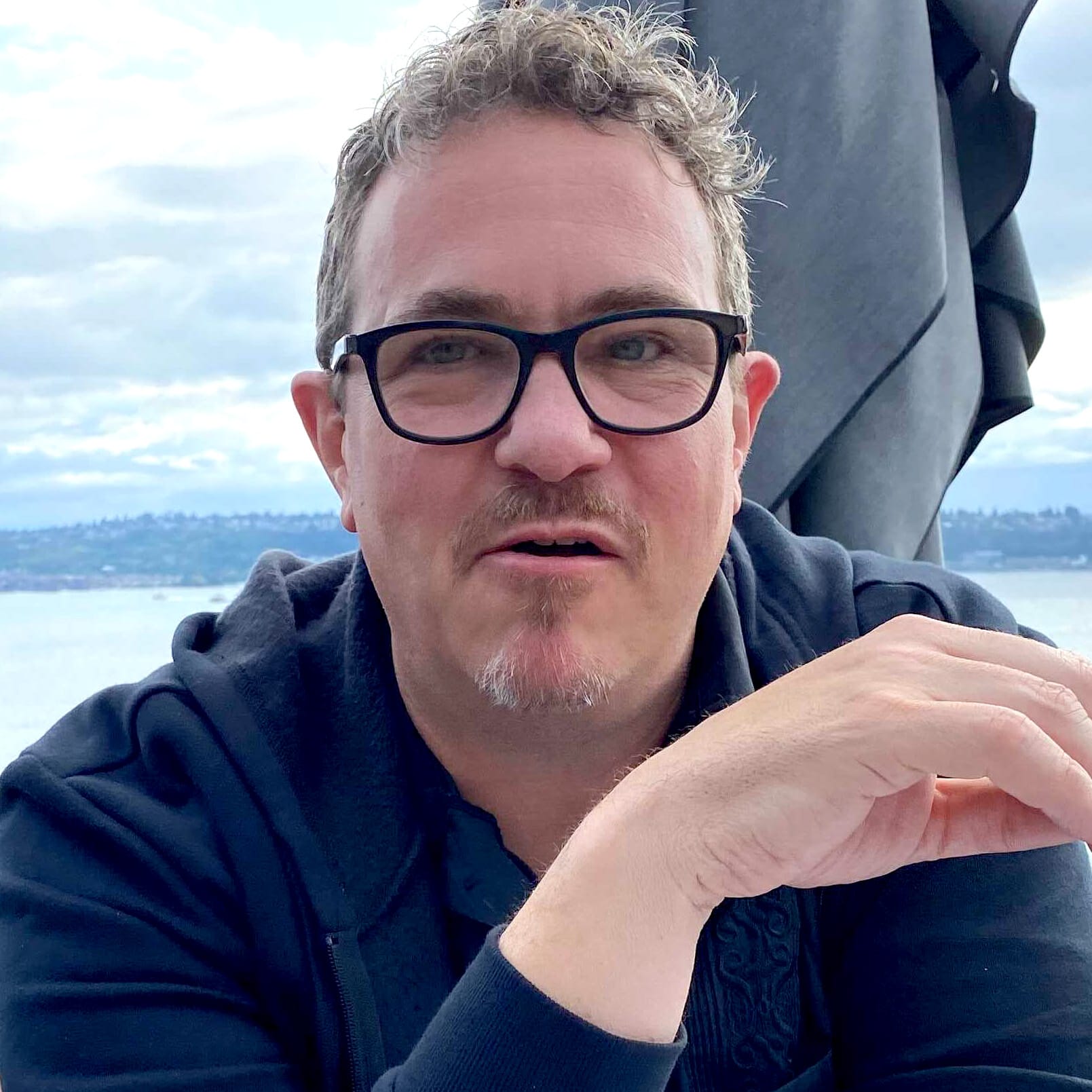 Picture of Bryan Zug in a black hoodie at a rooftop Happy Hour overlooking the Seattle Waterfront.