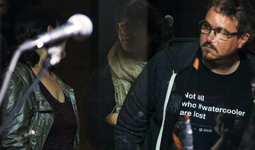 Bryan Zug in black hoodie and black t-shirt that reads "Not all who watercooler are lost". He is looking into the KEXP performance space where the musician Andrew Bird is playing.